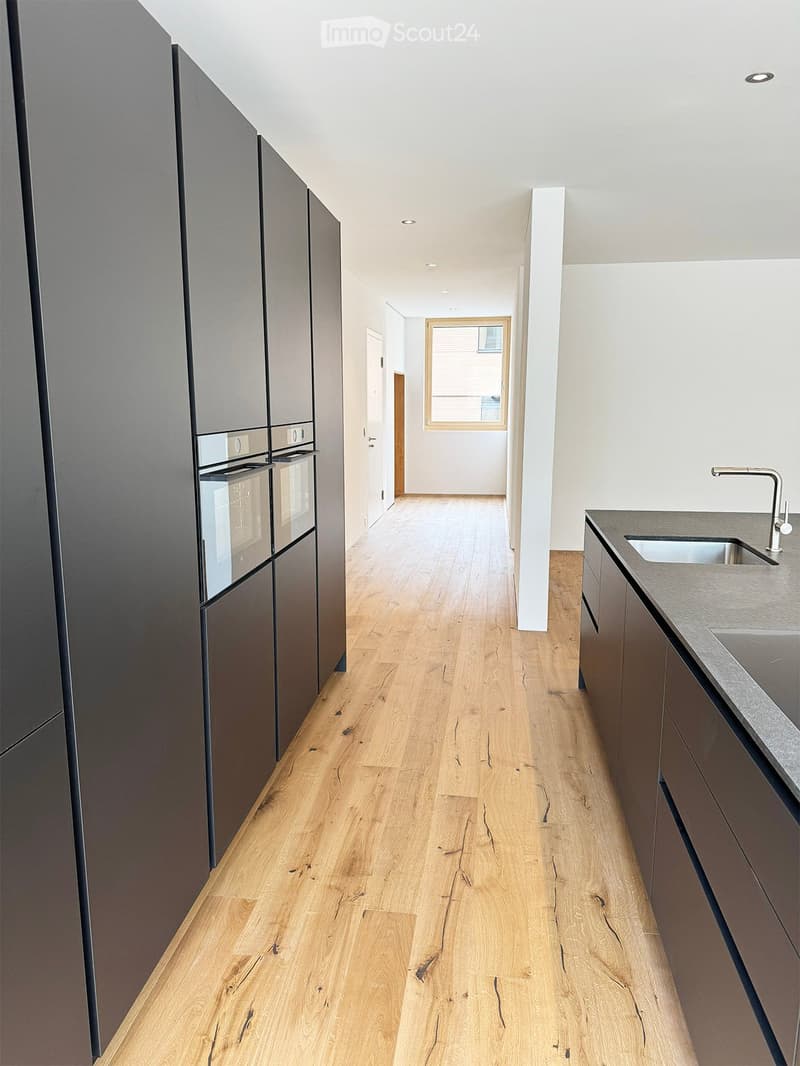 Modern hallway with light wooden flooring, grey cabinetry, and a window at the end. The space is clean and well-lit with recessed ceiling lights.
