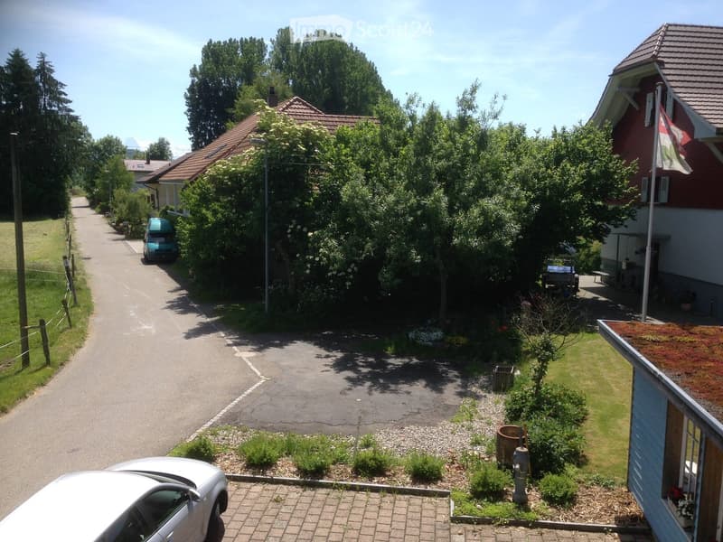 Residential street with houses, trees, and parked cars. Houses have red-tiled roofs and gardens.