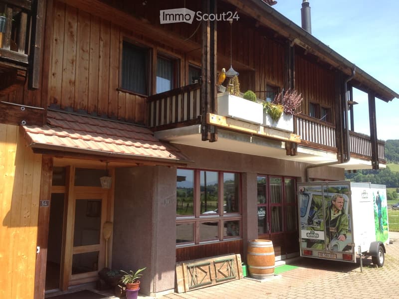 Two-story wooden building with a balcony, large windows, and a covered entrance area. A trailer is parked nearby, and there is a wooden barrel and potted plants on the ground.