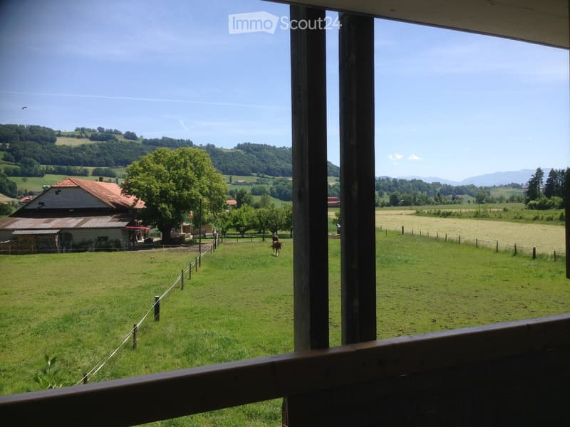 View from a balcony or terrace showing a green field, a small house, trees, and distant hills under a clear blue sky.