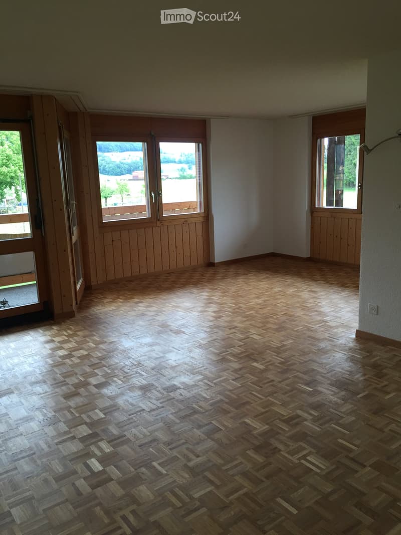 Empty room with wooden herringbone flooring, white walls, and large windows with wooden frames. The space is well-lit and has a neutral color scheme.