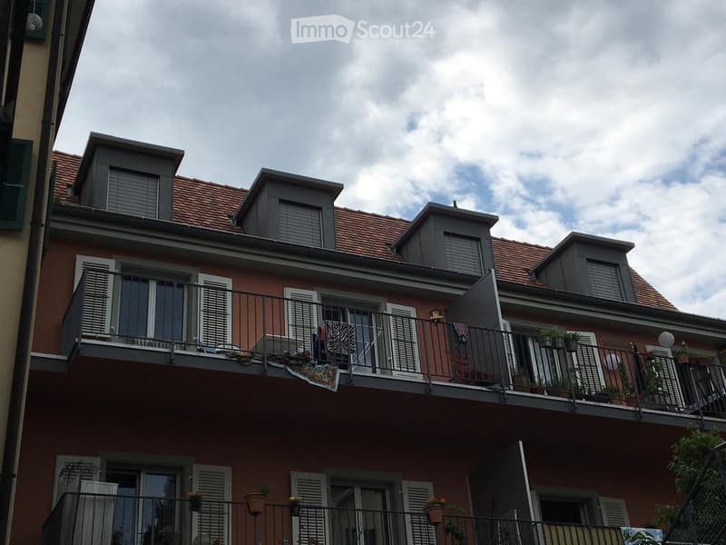 A multi-story residential building with red brick facade, multiple balconies, and potted plants. The roof features dormer windows with grey shutters.