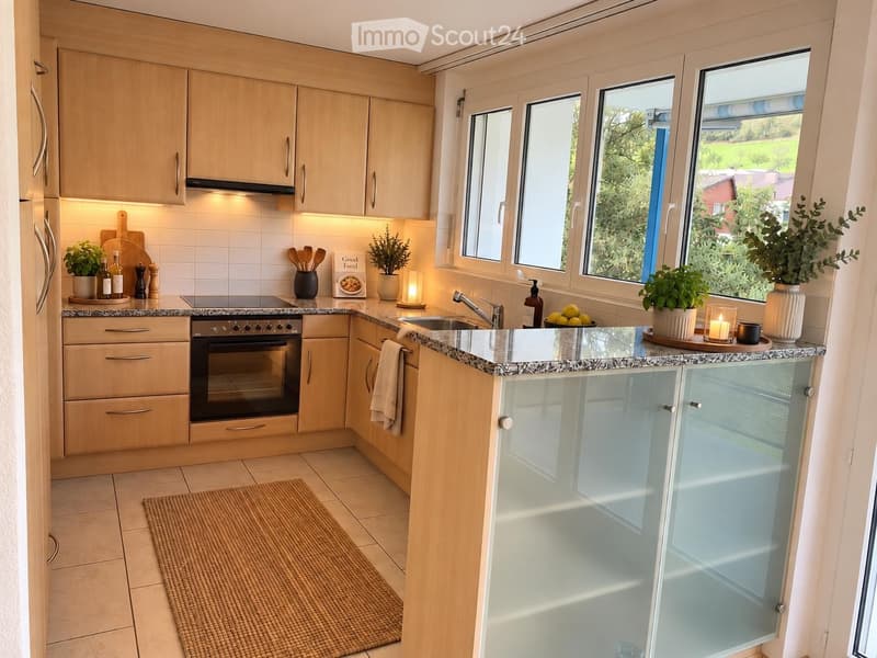 Modern kitchen with light wooden cabinets, granite countertops, stainless steel appliances, and large windows overlooking a green area.