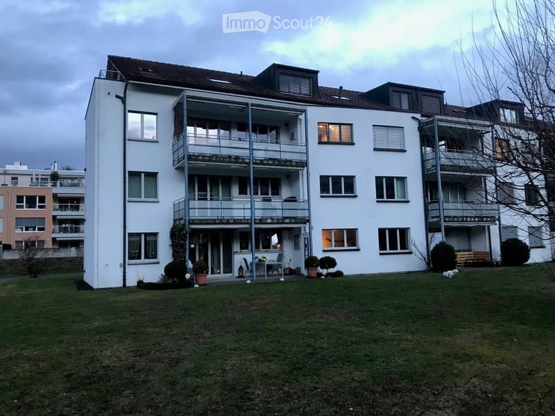 Modern white apartment building with multiple balconies, surrounded by a well-maintained lawn and small garden area.