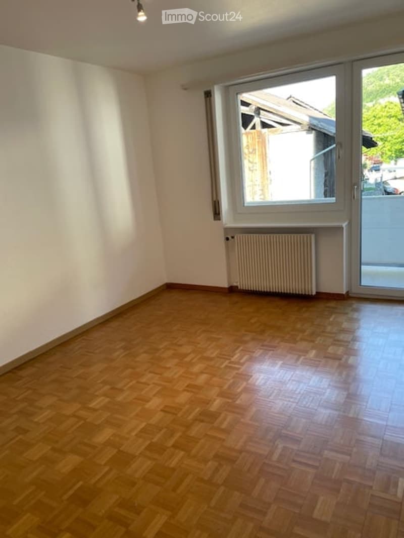 Empty room with wooden parquet flooring, white walls, a radiator, and a large window with a view of the outside.