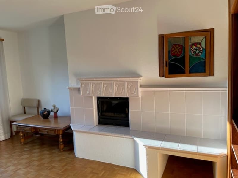 Living room with a white tiled fireplace, wooden floor, small table with a vase and statue, and a chair near the window.