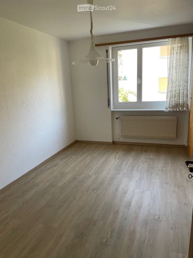 Empty room with wooden flooring, white walls, a window with curtains, and a radiator below the window.