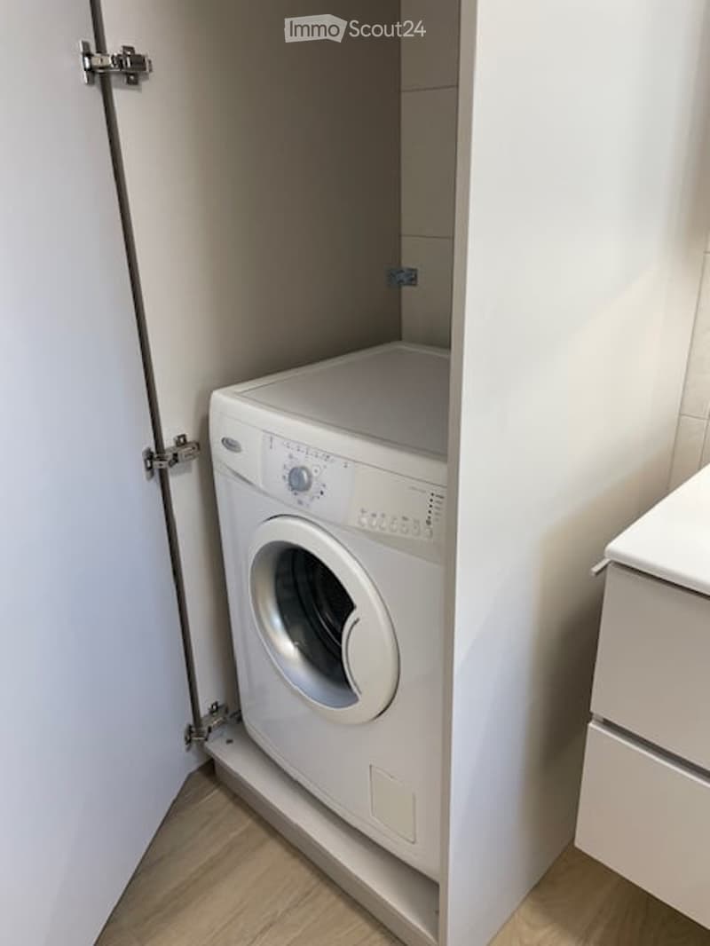 Compact bathroom with a white washing machine installed in a corner, white tiled walls, and wooden flooring.