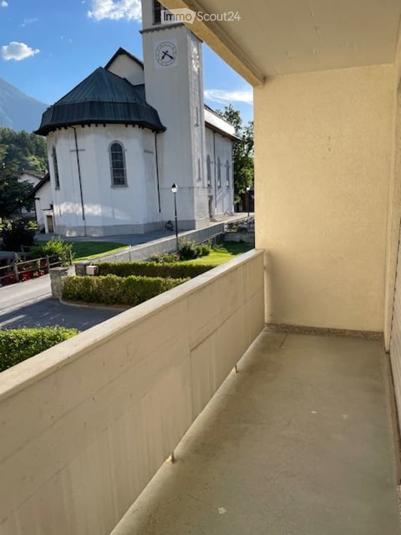 Balcony overlooking a white church with a clock tower, surrounded by greenery and a clear blue sky.