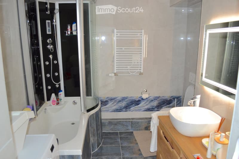 Modern bathroom with a bathtub, shower, sink, toilet, and a towel rack. The walls feature a marble-patterned design, and the floor is tiled. A wooden cabinet holds the sink and various toiletries.