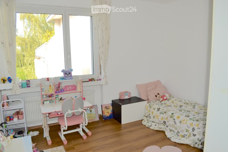 A well-lit bedroom with a single bed, a small desk with a chair, and various toys and storage items. The room features wooden flooring and large windows with curtains.