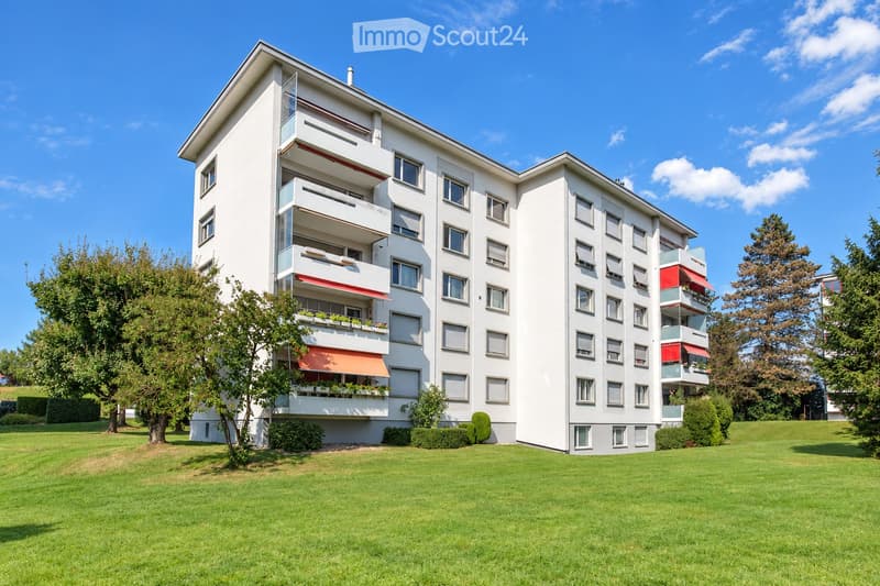 Modern multi-story apartment building with white facade, multiple balconies, and red awnings. Surrounded by green lawn and trees.