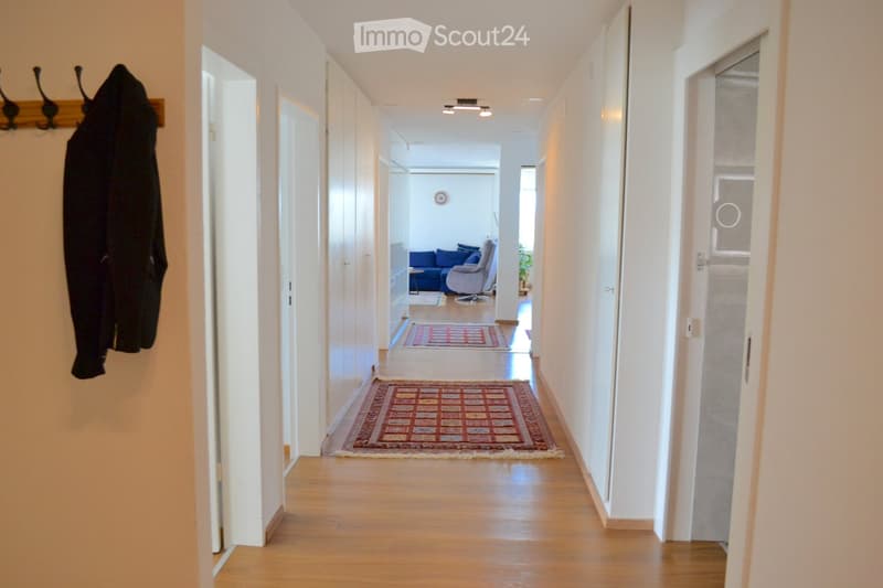 A long, well-lit hallway with wooden flooring, white walls, and two area rugs placed on the floor. A coat hangs on a rack on the left side, and a glimpse of a living room with a blue sofa and armchair is visible at the end of the hallway.