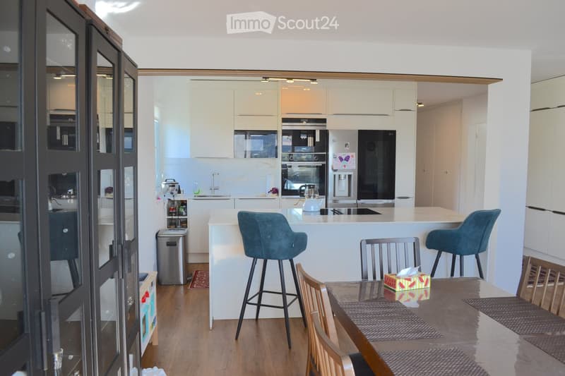 Modern kitchen with white cabinets, stainless steel appliances, and a central island with bar stools. Wooden dining table with chairs and a colorful watermelon-themed centerpiece.