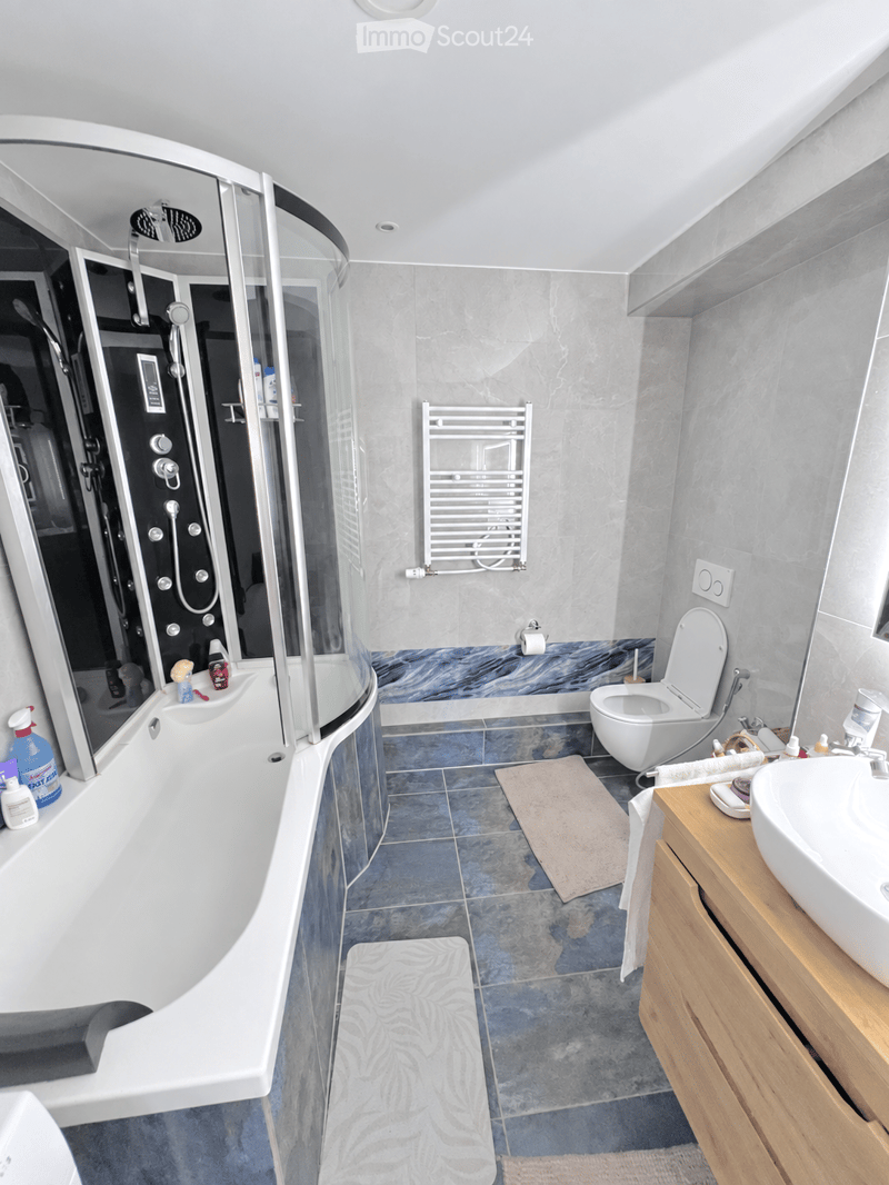 Modern bathroom with a bathtub, toilet, sink, and a glass-enclosed shower. Features a towel rack, wooden cabinet, and two bathroom mats on the floor.