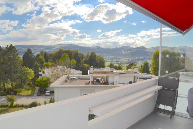 Balcony with a view of a suburban area, featuring a white railing, a single black wicker chair, and a scenic backdrop of trees, hills, and a partly cloudy sky.