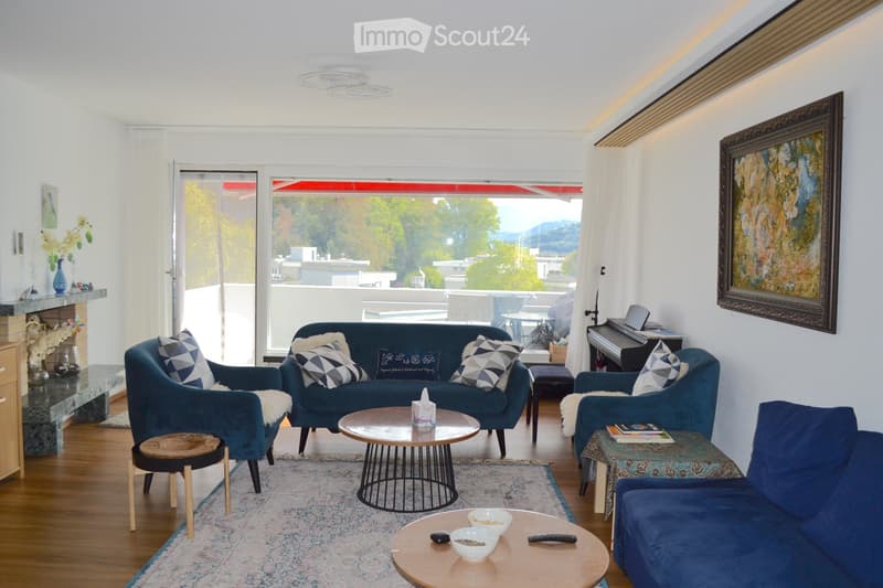 Modern living room with large windows offering a scenic view, featuring a blue sofa, armchairs, coffee tables, a piano, and a fireplace.
