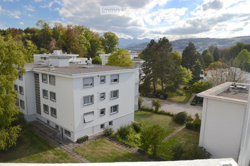 A modern white multi-story building with numerous windows, surrounded by greenery and trees, with a scenic mountain view in the background.