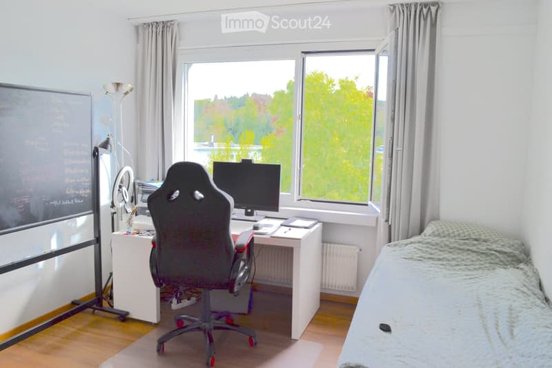 A furnished room with a bed, desk, chair, and large windows overlooking a green landscape. The room features a radiator, a mirror, and a curtain.