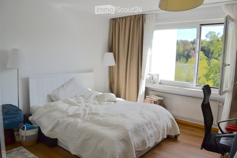 Cozy bedroom with a large bed, white bedding, wooden floor, large windows with curtains, and a view of greenery outside.