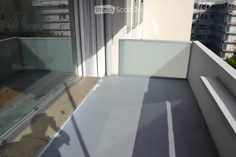 Modern balcony with concrete floor, glass railing, and a large window offering a view of the city.