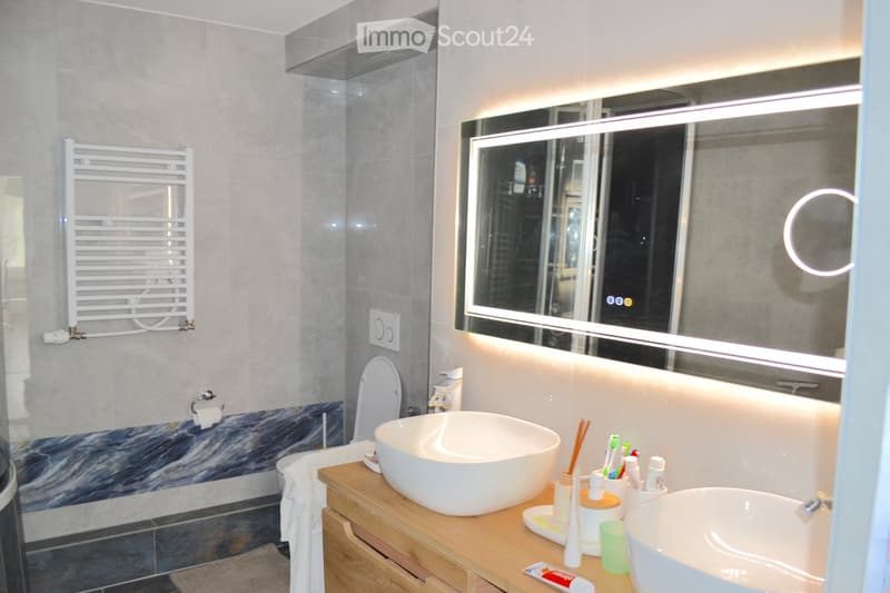 Modern bathroom with two sinks, a toilet, a large mirror with built-in lighting, a towel rack, and various toiletries on the countertop.