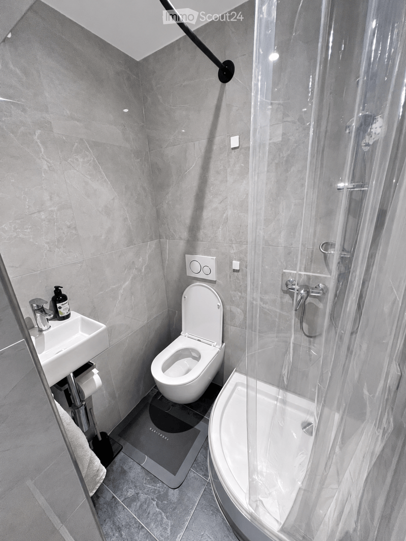 Modern bathroom with a toilet, sink, and walk-in shower with a glass enclosure. The walls and floor are covered in grey marble tiles, and a towel is draped over a silver towel rack.
