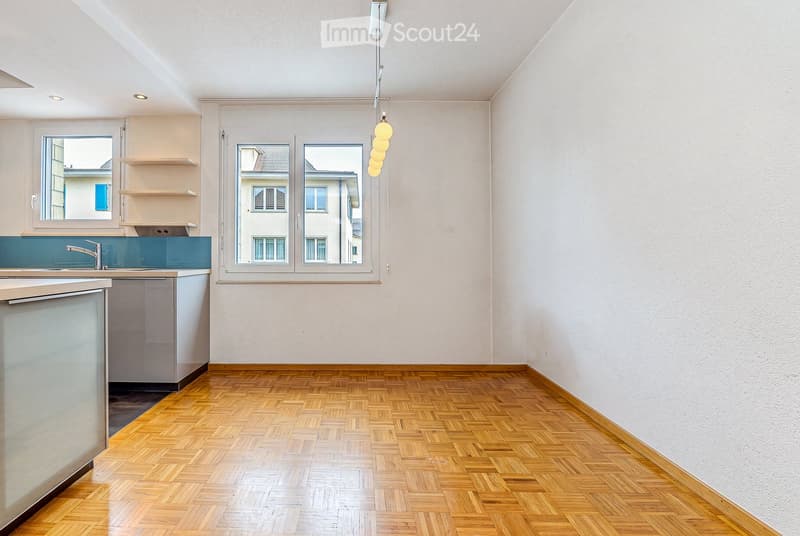 Empty kitchen with wooden herringbone flooring, white walls, and built-in cabinets. Features a window with a view of neighboring buildings, a hanging light fixture, and a sink with a modern faucet.