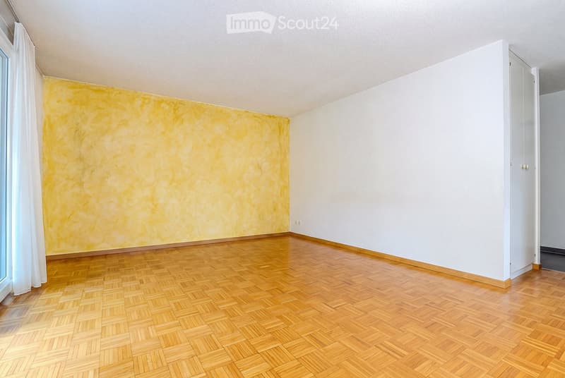 Empty room with wooden parquet flooring, white walls, and a large window with white curtains. The walls feature a textured yellow accent wall on one side.