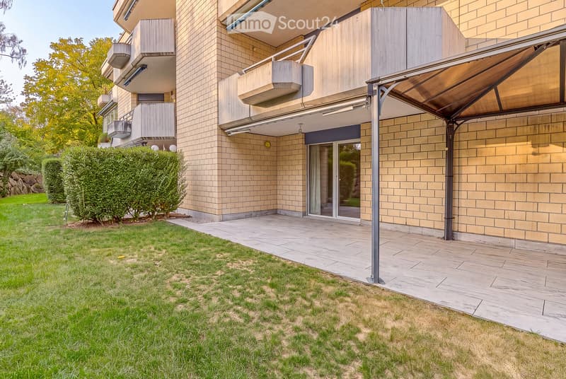 Modern multi-story building with brick and wood facade, balconies, and a covered entrance area. The surrounding area includes a well-maintained lawn and paved walkway.