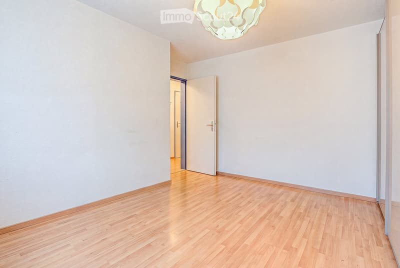 Empty room with light-colored walls and wooden flooring, featuring a ceiling light fixture and an open doorway leading to another room.