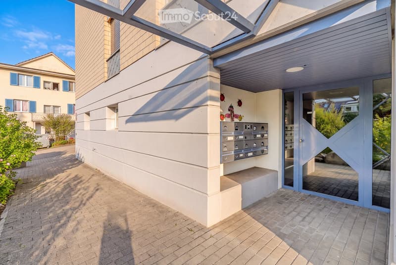 Modern building entrance with mailboxes, glass door, and decorative wall art. The entrance features a covered area with a sleek design and a paved walkway.