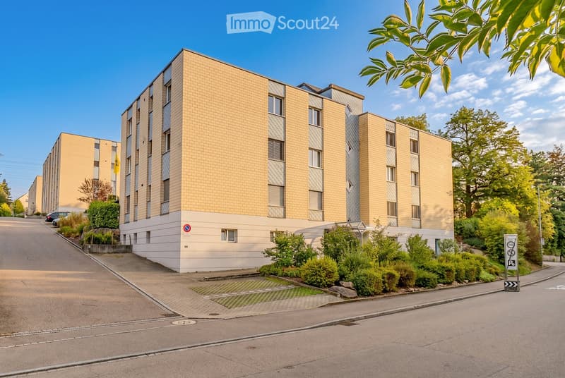Modern multi-story residential building with beige brick facade, large windows, and surrounding greenery.
