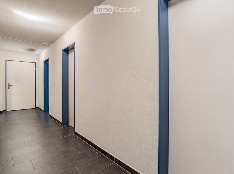 Empty hallway with tiled floor, white walls, and blue accent walls. Features two doors and a light switch on the wall.