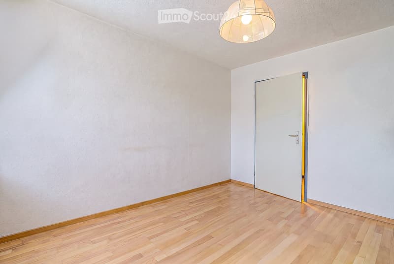 Empty room with light-colored walls, wooden flooring, and a single door leading to another space. A modern pendant light hangs from the ceiling.