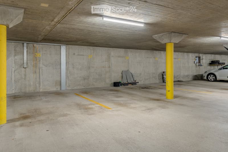 Underground parking area with concrete walls, floor markings, and a few parked cars. Yellow pillars and numbered spaces are visible.