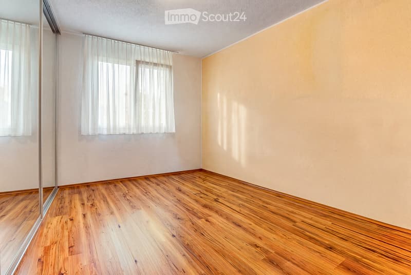 Empty room with wooden flooring, white walls, and large windows with sheer curtains allowing natural light to enter.