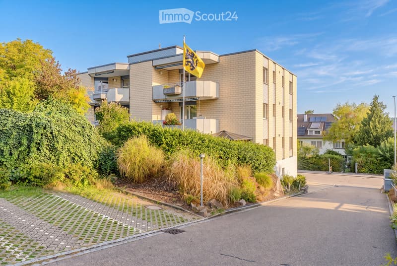 Modern multi-story apartment building with balconies, surrounded by greenery and a paved driveway.