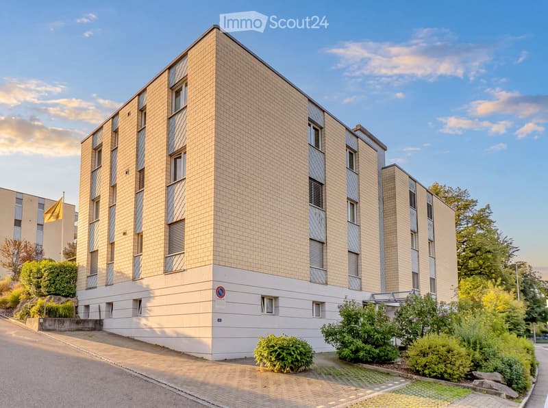 Modern multi-story residential building with beige brick facade, large windows, and greenery around the base.