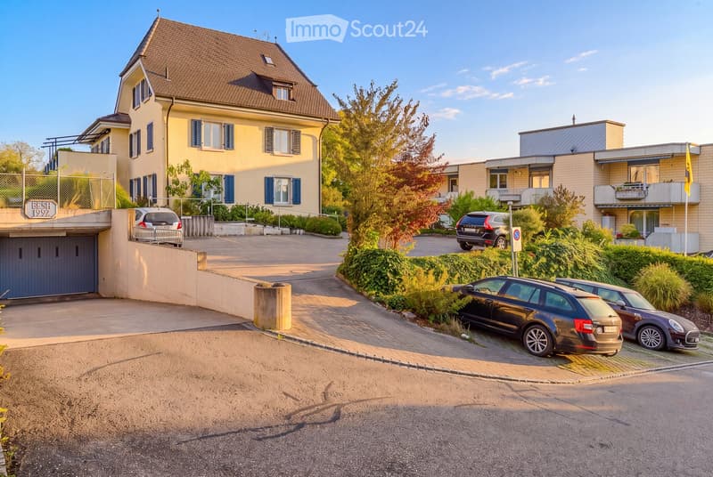 Two-story residential building with a garage, surrounded by greenery and parked cars.