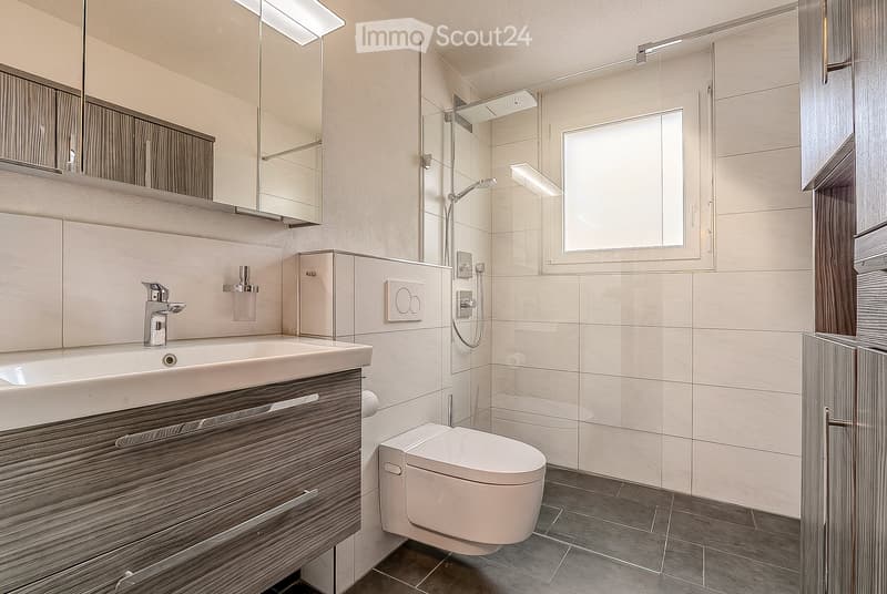 Modern bathroom with a white sink, sleek faucet, large mirror, toilet, and a glass-enclosed shower with a modern showerhead. The walls are tiled in a light, neutral color, and the floor is dark grey.