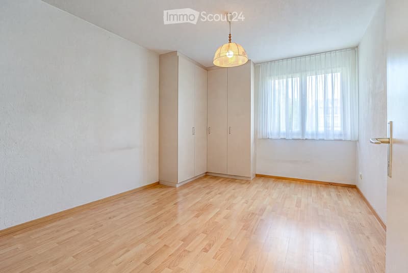 Empty room with light wooden flooring, white walls, built-in wardrobes, a window with white curtains, and a hanging light fixture.