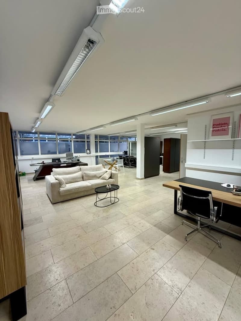 Bright, open-plan room with large windows, a cream-colored sofa, a coffee table, a desk with an office chair, and shelves with books and decor.