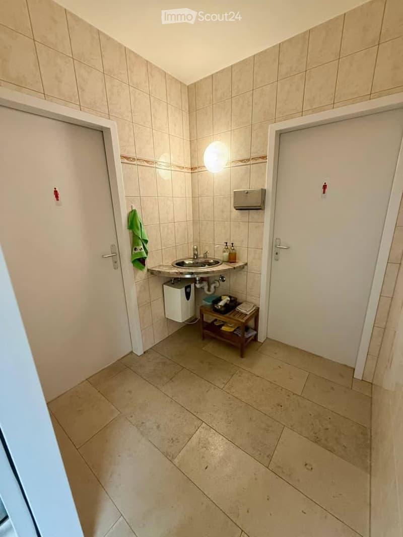 A small bathroom with beige tiled walls and floor, a sink with a mirror above it, a towel hanging on the wall, and a small shelf holding various items.
