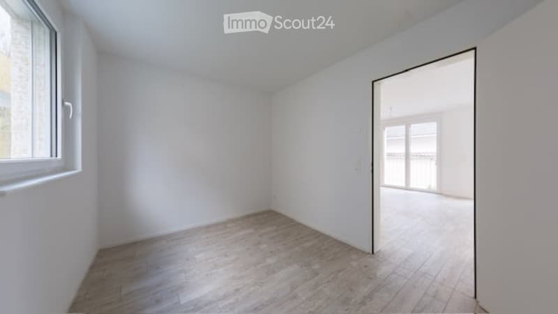 Empty room with wooden flooring, white walls, and a large window. An open doorway leads to a balcony area with glass railings.