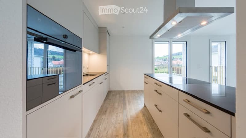 Modern kitchen with white cabinets, stainless steel oven, countertop, and wooden flooring. Large windows offer a scenic view.