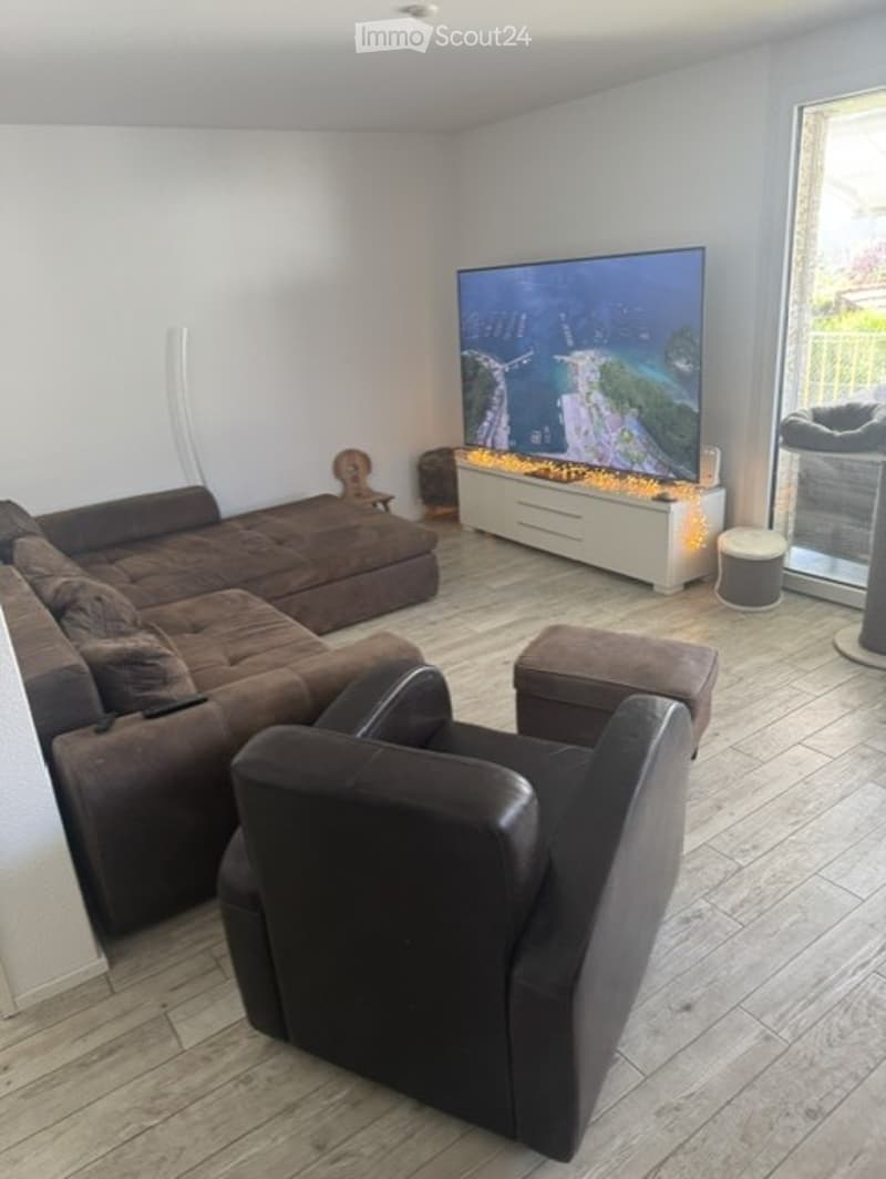 Modern living room with large windows offering a scenic view, featuring a brown sectional sofa, matching armchair, ottoman, TV stand with a flat-screen TV, table lamp, and a small stool.