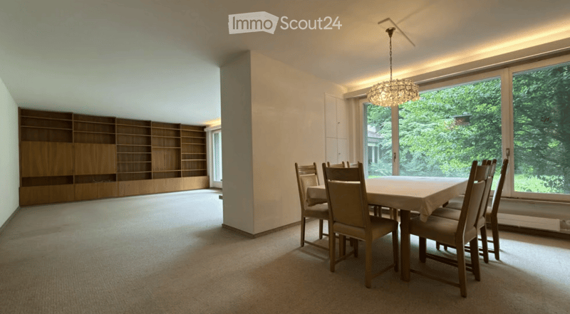 Bright dining room with large windows, a wooden dining table surrounded by chairs, and a tall wooden bookshelf against the wall.