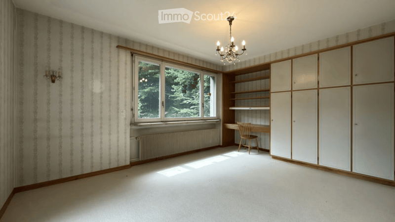 Empty room with white walls, wooden trim, large window, built-in shelving unit, and a small desk with a chair.