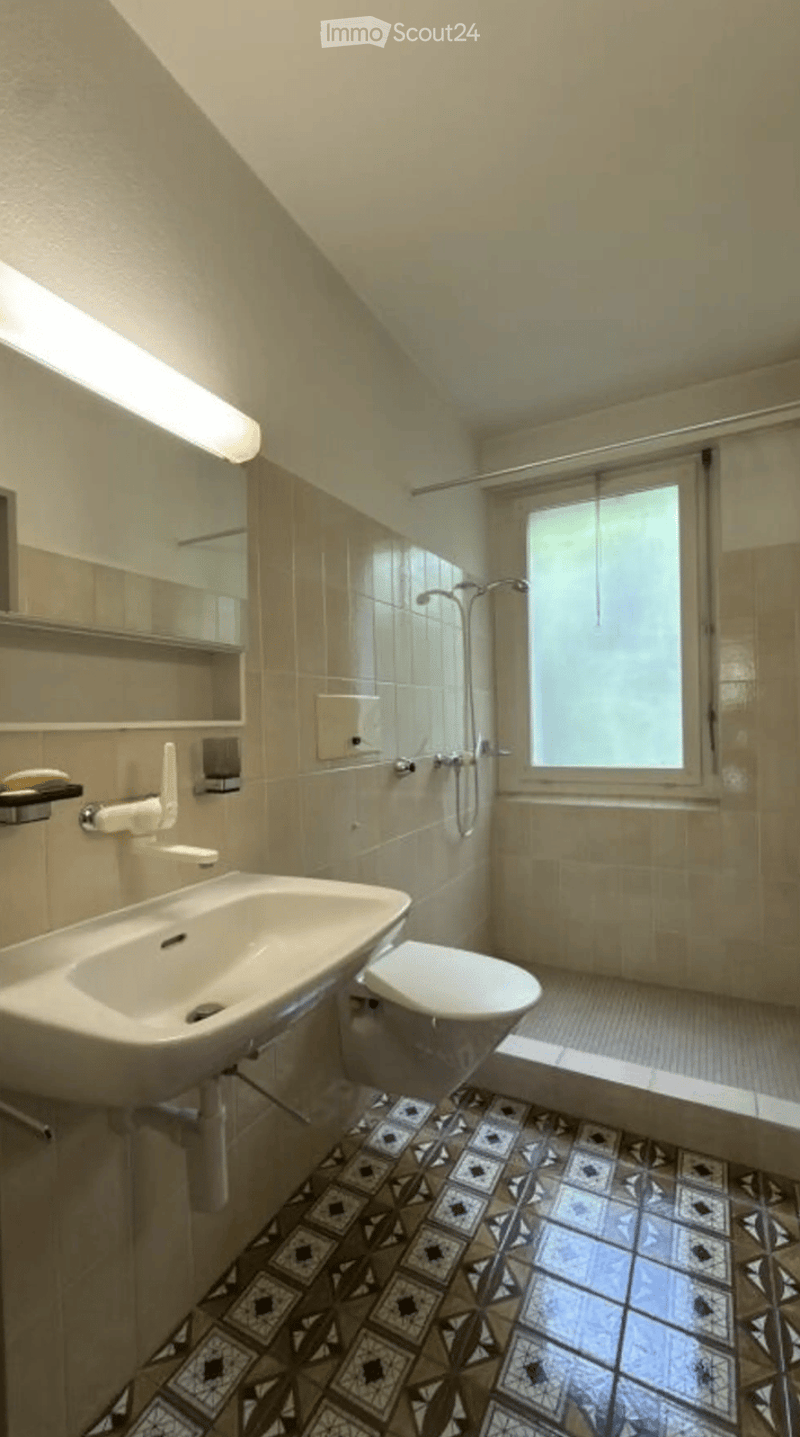 Bathroom with a sink, toilet, shower, mirror, and light fixture. The floor features a patterned tile design, and the walls are tiled with beige tiles.